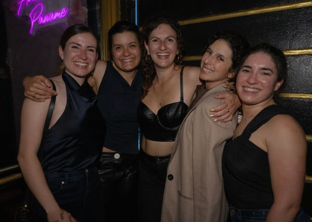 Five smiling women arm-in-arm posing in a dim bar with a purple neon sign — lively city nightlife group photo.
