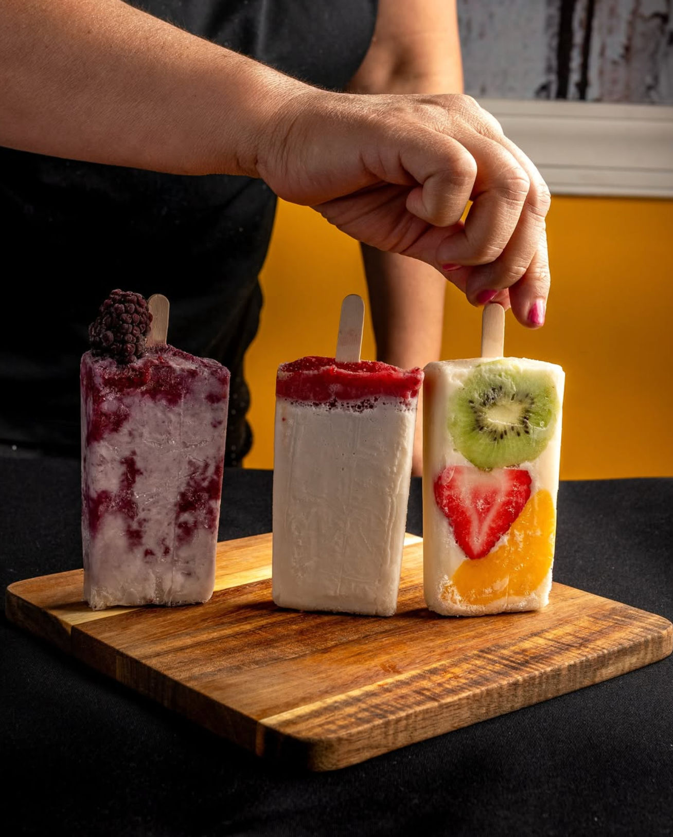 Hand grabbing a fruit-filled popsicle from three colorful gourmet popsicles on a wooden board — blackberry swirl, creamy raspberry-topped, and kiwi‑strawberry‑mango slice pop against a bright yellow background.