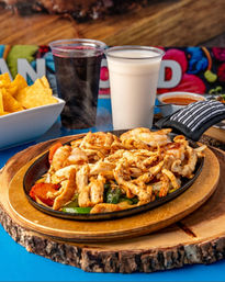 Steaming Mexican-style chicken and shrimp fajitas with sautéed bell peppers and onions on a sizzling cast-iron skillet, served on a wooden board with tortilla chips, salsa and two drinks (creamy horchata and dark hibiscus agua fresca).