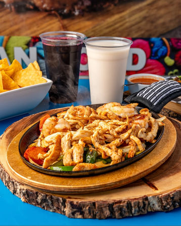 Steaming Mexican-style chicken and shrimp fajitas with sautéed bell peppers and onions on a sizzling cast-iron skillet, served on a wooden board with tortilla chips, salsa and two drinks (creamy horchata and dark hibiscus agua fresca).
