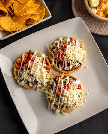 Three colorful Mexican sopes on a white square plate topped with seasoned meat, shredded lettuce, diced tomatoes, crumbled queso fresco and drizzled crema, with a bowl of tortilla chips and corn salad nearby