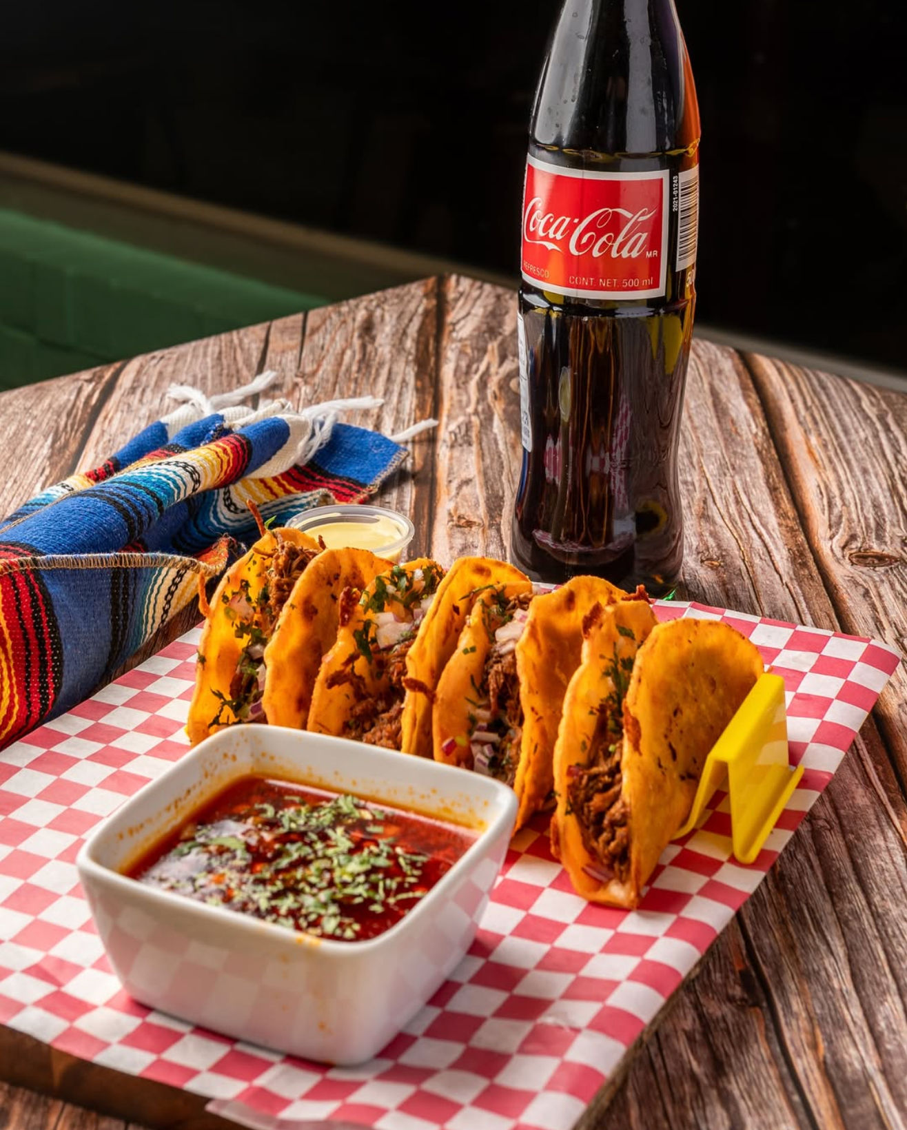 Golden birria tacos in a rack with a bowl of red consomé for dipping, crema cup and a cold soda bottle on a rustic wooden table beside a colorful Mexican blanket