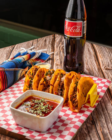 Golden birria tacos in a rack with a bowl of red consomé for dipping, crema cup and a cold soda bottle on a rustic wooden table beside a colorful Mexican blanket