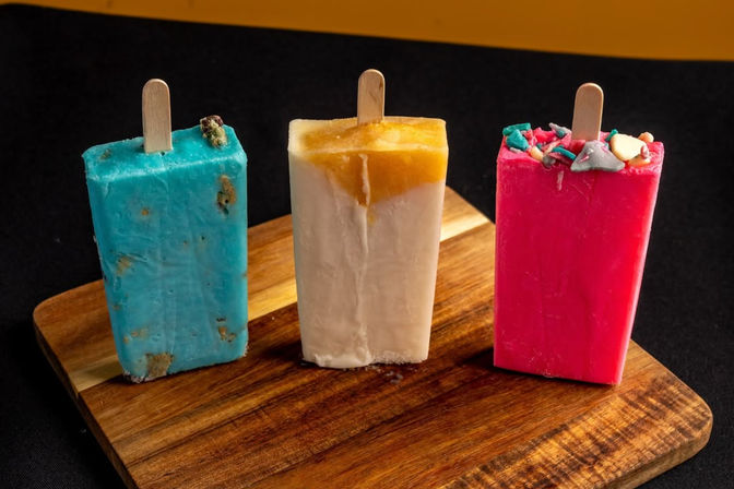 Three colorful rectangular popsicles—turquoise with cookie bits, creamy white with a yellow mango swirl, and bright pink topped with rainbow sprinkles—standing on a wooden serving board against a black background.
