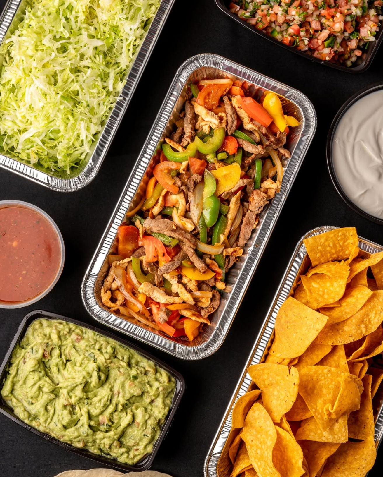 Aluminum catering trays with sizzling beef and chicken fajitas with colorful bell peppers and onions, surrounded by tortilla chips, creamy guacamole, pico de gallo, salsa, shredded lettuce and sour cream on a black backdrop.