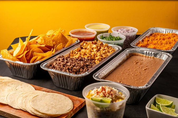 Vibrant Mexican taco bar catering spread with soft corn tortillas, tortilla chips, grilled beef and chicken, refried beans, Mexican rice, salsas, chopped cilantro and onions, lime wedges and elote-style corn in foil trays.