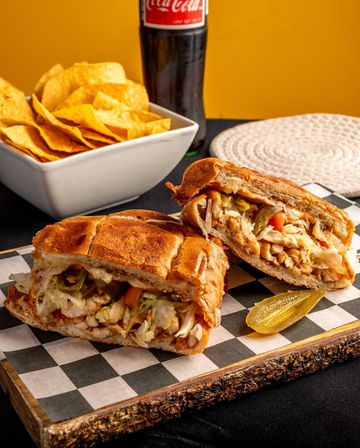 Mexican-style grilled chicken torta cut in half on checkered paper atop a wooden board, filled with shredded lettuce, pickled jalapeños and tomatoes, served with a pickle spear, a bowl of tortilla chips and a glass soda bottle against a warm yellow backdrop.