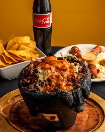 Mexican molcajete overflowing with melted cheese, grilled meats and peppers on a wooden board, served with tortilla chips, bacon-wrapped jalapeños and dipping sauce, plus a glass bottle of cola against a warm yellow backdrop.