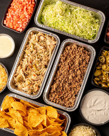 Party-ready Mexican-style taco bar catering spread in foil trays with shredded chicken, seasoned ground beef, shredded lettuce, diced tomatoes, jalapeños, tortilla chips, shredded cheese and bowls of sour cream and queso.