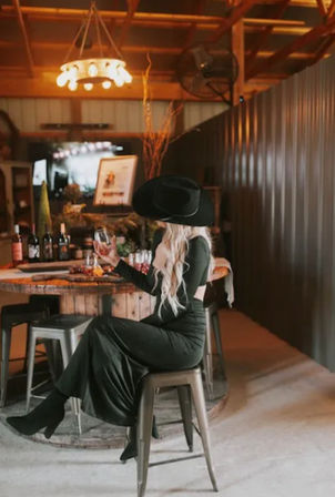 Country-chic scene: blonde woman in a black cowboy hat and long black dress sits on a metal stool sipping wine in a rustic indoor tasting room with a barrel table, bottles, and warm pendant lights.