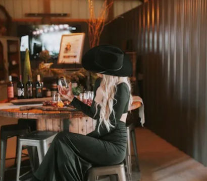 Country-chic scene: blonde woman in a black cowboy hat and long black dress sits on a metal stool sipping wine in a rustic indoor tasting room with a barrel table, bottles, and warm pendant lights.