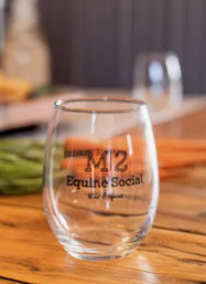 Stemless wine glass with an equine-themed logo on a rustic wooden bar counter, colorful napkins and blurred bar stools in the background.