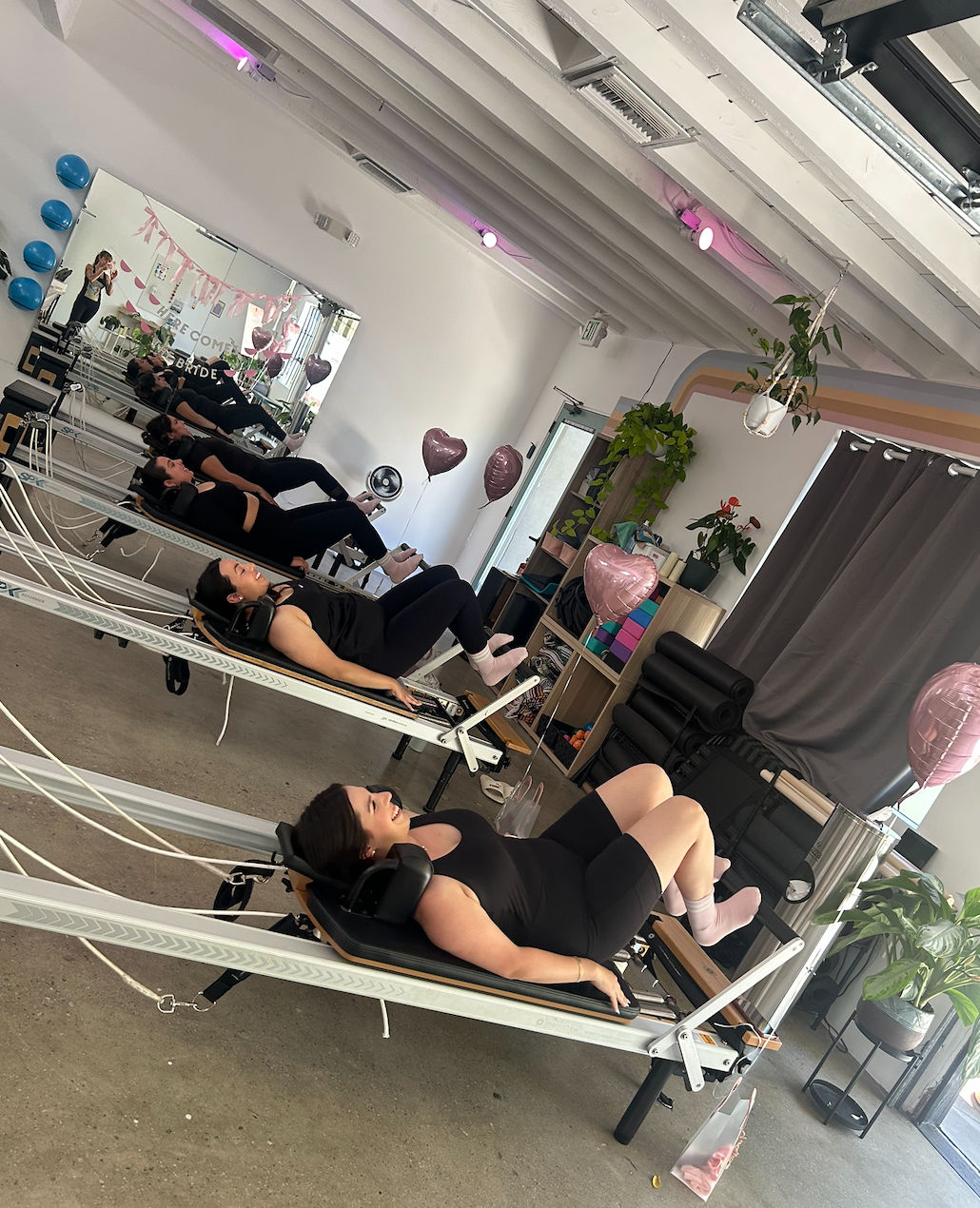 Group pilates reformer class with women in black workout gear on reformer machines in a bright fitness studio with mirrored wall, pink heart-shaped balloons, hanging plants and natural light.