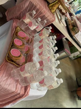 Pink-themed event refreshment table with rows of bottled water wearing custom pink labels and a gold-trimmed tray of individually wrapped cookies on a crinkled pink table runner.