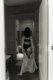 Black-and-white doorway view of a woman seen from behind in a black bikini walking into a wooden indoor spa sauna while holding a patterned towel.
