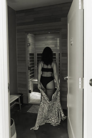 Black-and-white doorway view of a woman seen from behind in a black bikini walking into a wooden indoor spa sauna while holding a patterned towel.