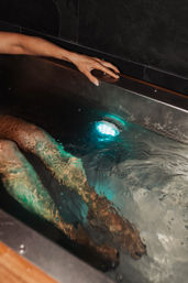 Person soaking in an indoor spa tub, legs submerged in rippling water lit by a teal underwater LED and a hand resting on the metal rim.