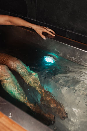 Person soaking in an indoor spa tub, legs submerged in rippling water lit by a teal underwater LED and a hand resting on the metal rim.