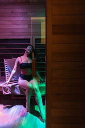 Woman in a black bikini relaxing on a towel-covered bench inside a modern wooden sauna, bathed in purple and green mood lighting.
