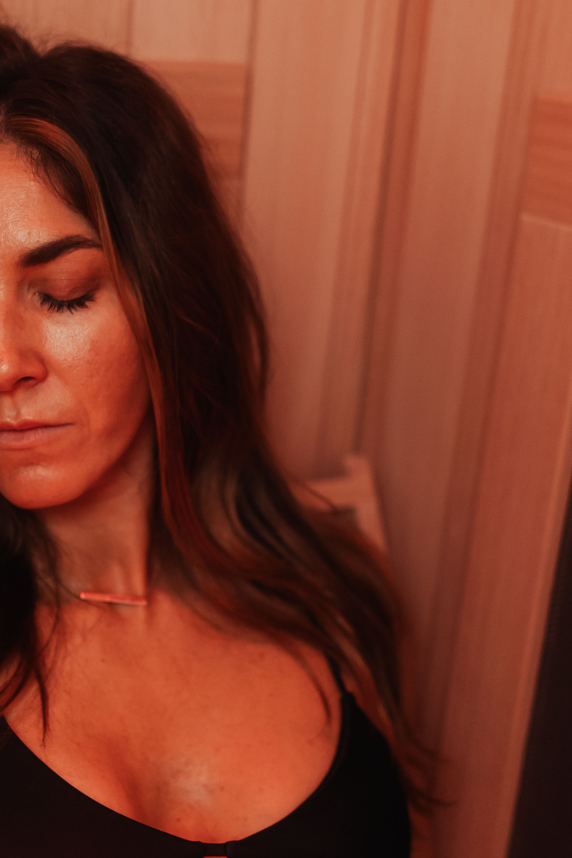 Close-up portrait of a relaxed woman with eyes closed, long hair, black top and bar necklace, leaning against wooden-paneled wall in warm red ambient light — spa-style relaxation.