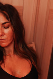 Close-up portrait of a relaxed woman with eyes closed, long hair, black top and bar necklace, leaning against wooden-paneled wall in warm red ambient light — spa-style relaxation.