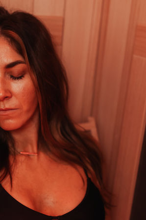 Close-up portrait of a relaxed woman with eyes closed, long hair, black top and bar necklace, leaning against wooden-paneled wall in warm red ambient light — spa-style relaxation.