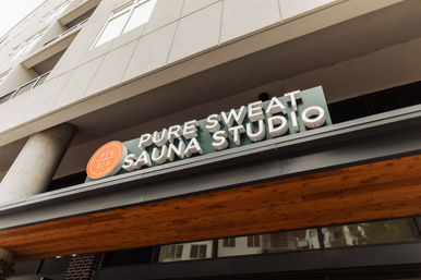 Angled shot of a modern urban sauna studio storefront sign — bold 3D letters, orange circular logo, and warm wood canopy.