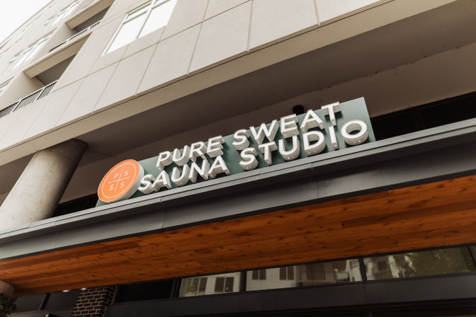 Angled shot of a modern urban sauna studio storefront sign — bold 3D letters, orange circular logo, and warm wood canopy.