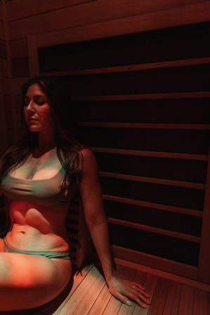 Woman in a bikini relaxing on a wooden bench inside a dim, red-lit infrared sauna — serene spa wellness scene.