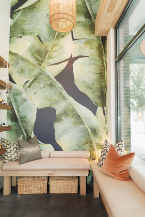 Cozy storefront seating nook with oversized green banana-leaf mural, pale wood benches, patterned and velvet pillows, rattan pendant light and wicker storage baskets