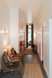 Cozy modern hallway with curved rattan lounge chairs and lamp, jute runners on a dark floor, bright red doors along the corridor and a curtained window at the far end.
