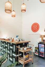 Bright sauna studio reception with woven pendant lamps, wood-top desk with geometric tile base, rattan cart stacked with books and candles, mini fridge with beverages, and a round orange wall logo.