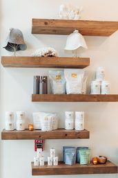 Rustic wooden floating shelves on a white wall holding wellness and skincare products—white jars, resealable pouches and packets, candles, coral decor and felt hats in a cozy spa display.