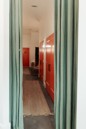 Modern interior hallway framed by green curtains, featuring bold red paneled doors with black handles, a woven runner rug on a concrete floor and warm wall sconce lighting.