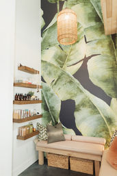 Cozy spa-style seating nook with large green banana-leaf mural, woven pendant light, wooden shelves of skincare bottles, cushioned bench and wicker storage baskets