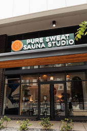 Urban sauna studio storefront with green sign and orange round logo, glass doors, warm hanging lights and sidewalk plants