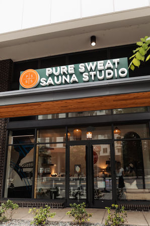 Urban sauna studio storefront with green sign and orange round logo, glass doors, warm hanging lights and sidewalk plants