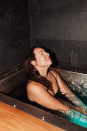 Woman in a black swimsuit relaxing with eyes closed in a blue-lit plunge tub against dark stone spa walls