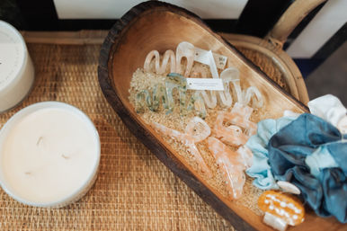 Pastel marbled acrylic hair clips and blue fabric scrunchies arranged on rice inside a rustic wooden dough bowl on a woven tabletop next to a white candle