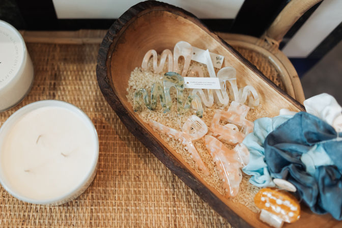 Pastel marbled acrylic hair clips and blue fabric scrunchies arranged on rice inside a rustic wooden dough bowl on a woven tabletop next to a white candle