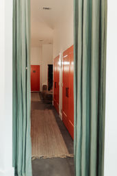 Interior hallway framed by green curtains, featuring bright red doors with black handles, a natural-fiber runner on a concrete floor, and warm wall sconce lighting