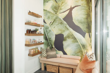 Cozy spa relaxation nook with oversized tropical leaf mural, wooden floating shelves displaying skincare bottles and jars, blush-cushioned bench with decorative pillows and woven storage baskets.
