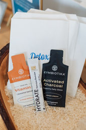 Three detox supplement sachets — a vitamin liposomal pouch, an activated charcoal pouch, and an electrolyte hydrate stick — nestled in rice inside a wooden tray with a 'Detox' card behind, spa-wellness display.