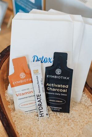Three detox supplement sachets — a vitamin liposomal pouch, an activated charcoal pouch, and an electrolyte hydrate stick — nestled in rice inside a wooden tray with a 'Detox' card behind, spa-wellness display.