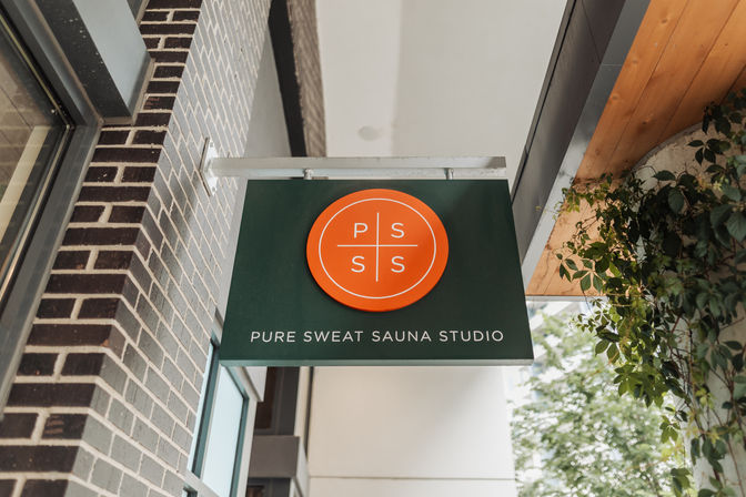 Green hanging storefront sign with orange circular logo and white text reading 'sauna studio' on a brick urban facade, framed by a wooden overhang and nearby greenery