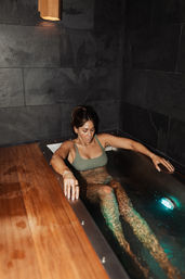 Woman in a green swimsuit relaxing in a narrow indoor spa plunge tub with warm wood trim, teal underwater light and dark slate tile walls