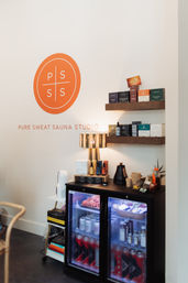 Bright sauna studio reception interior with an orange circular logo on a white wall, wooden shelves displaying wellness products, a lit table lamp, and a glass-front mini fridge stocked with bottled drinks and rolled towels — cozy, modern wellness vibe.