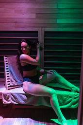 Person in black swimwear reclining on a wooden sauna bench with a towel, bathed in pink and green mood lighting for spa-style relaxation.