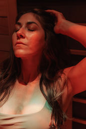 Close-up of a woman relaxing in a wood-paneled sauna under warm red light, eyes closed and hand resting behind her head, enjoying a calm spa moment.