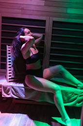Woman in black swimwear reclining on a wood-paneled sauna bench with a towel, bathed in magenta and green LED light — relaxing wellness scene.